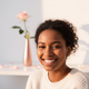 Headshot of a smiling young woman of color with a soft, sunlit glow and a blurred blush pink and white background.