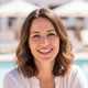 A happy woman in her late 20s to early 30s smiling warmly, with a blurred background hinting at a sunny outdoor setting.